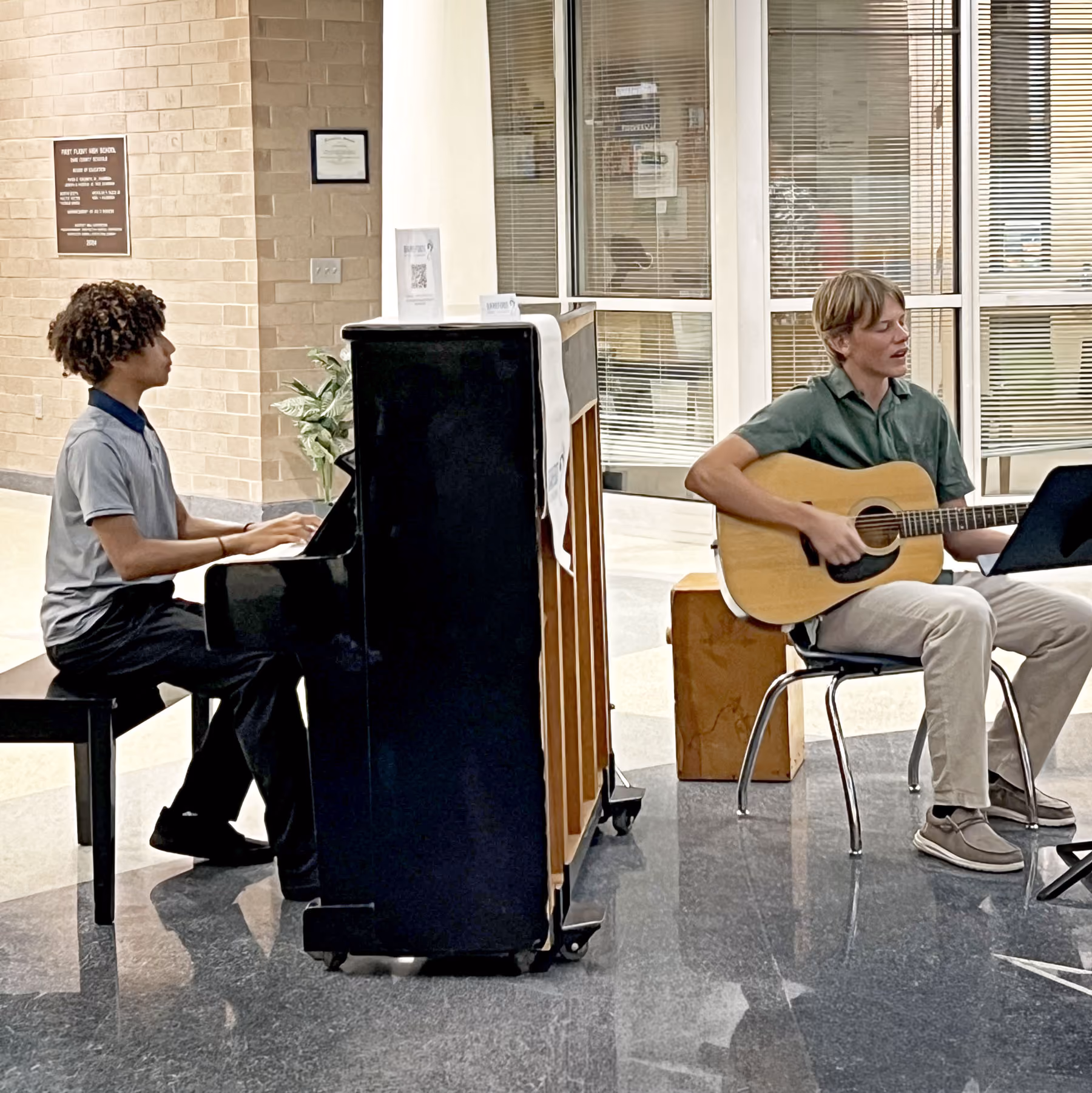 Two young men performing music indoors, one playing an upright piano and the other playing an acoustic guitar while sitting on chairs, with glass doors and windows in the background.