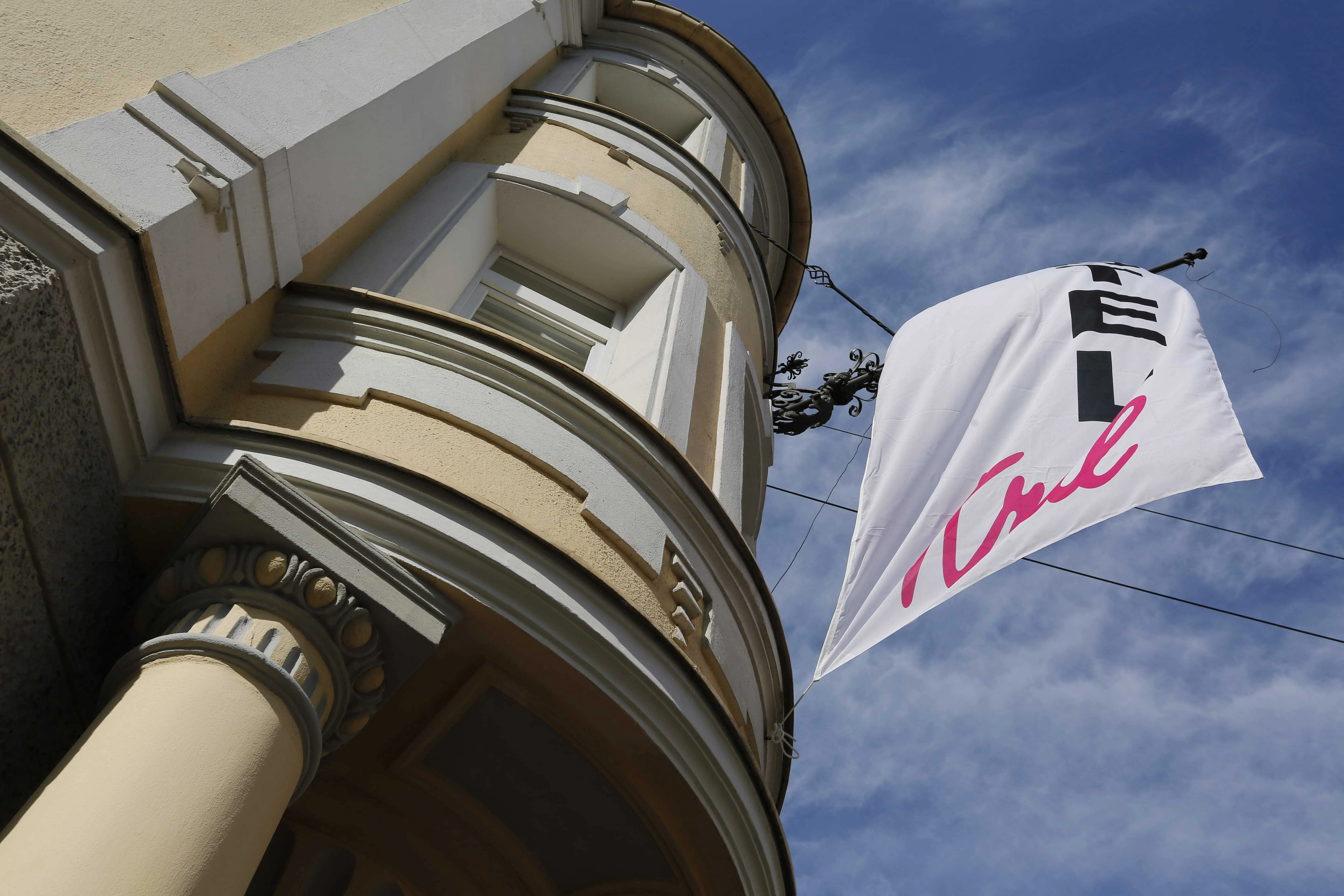Blick nach oben auf ein historisches Gebäude mit Säule und Rundbogenfenster, an dem eine weiße Bannerfahne mit schwarzer und pinker Schrift im Wind weht, vor blauem Himmel mit dünnen Wolken.