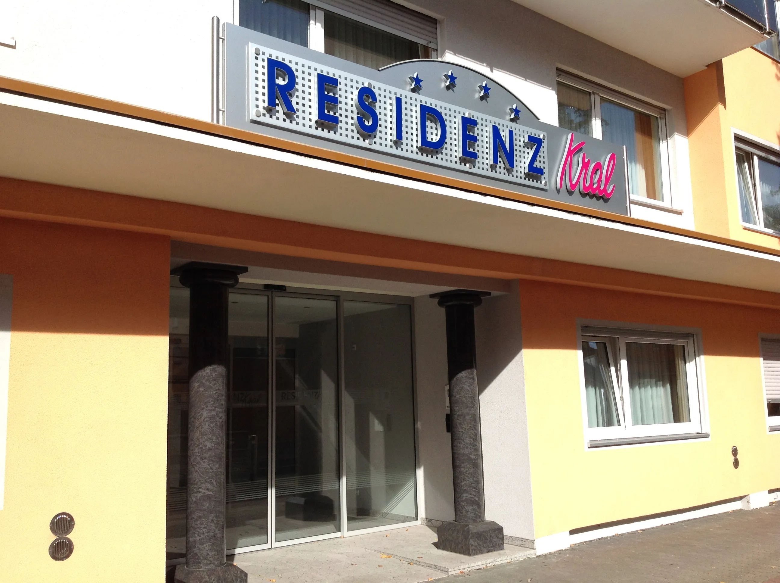 Eingang eines Gebäudes mit zwei schwarzen Marmorsäulen und einem Schild mit der Aufschrift 'RESIDENZ Kral' in blauen und roten Buchstaben.
