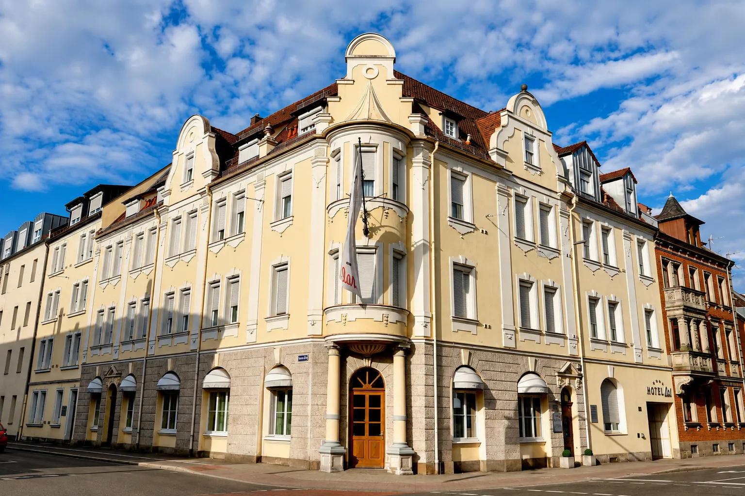 Eckgebäude eines dreistöckigen Hotels mit beiger Fassade, Säulen am Eingang, mehreren Fenstern und einem Fahnenmast, unter blauem Himmel mit Wolken.