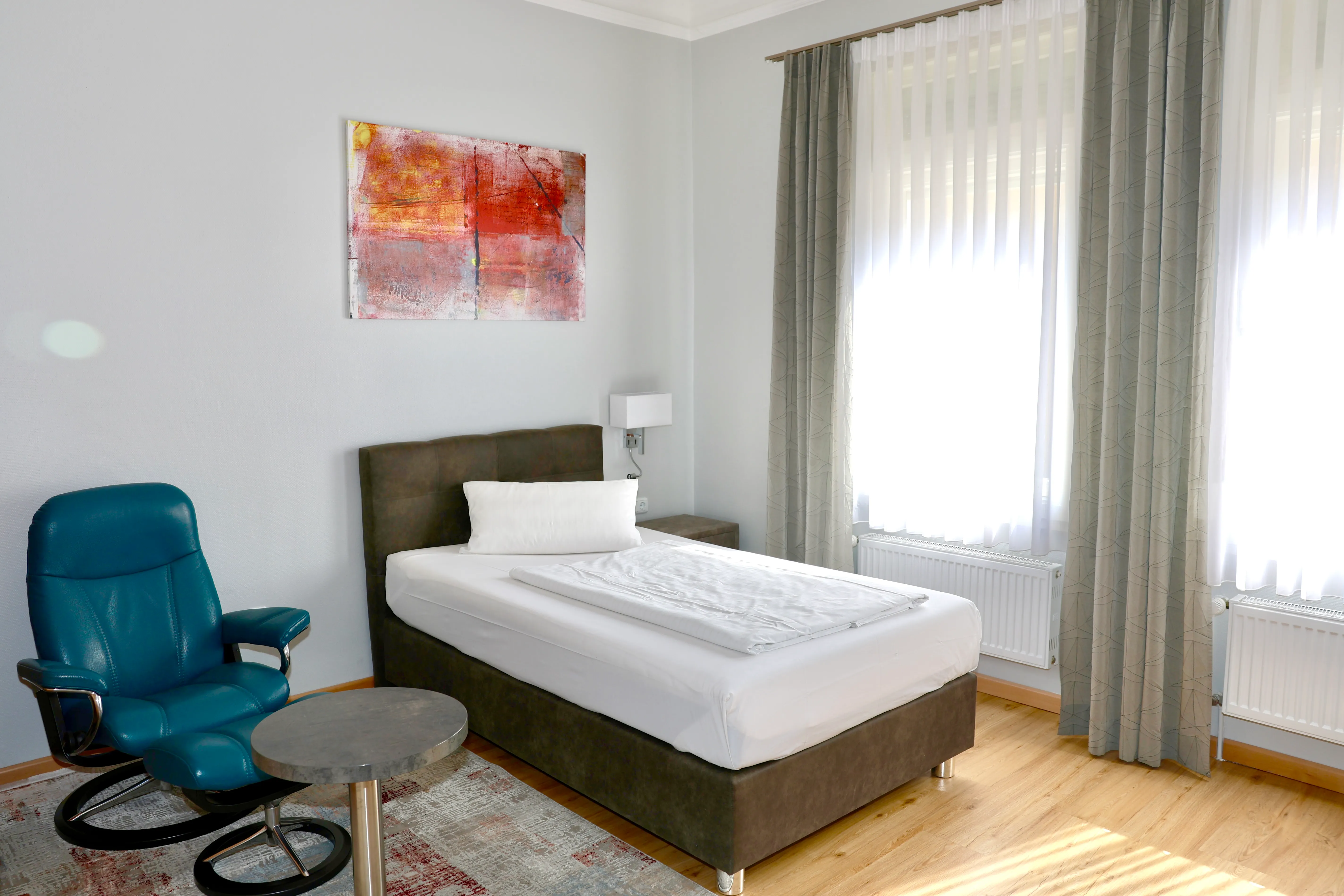 Helles Zimmer mit Einzelbett mit weißer Bettwäsche, blauem Ledersessel, kleinem runden Tisch, gemustertem Teppich und großem Fenster mit halbtransparenten Vorhängen.