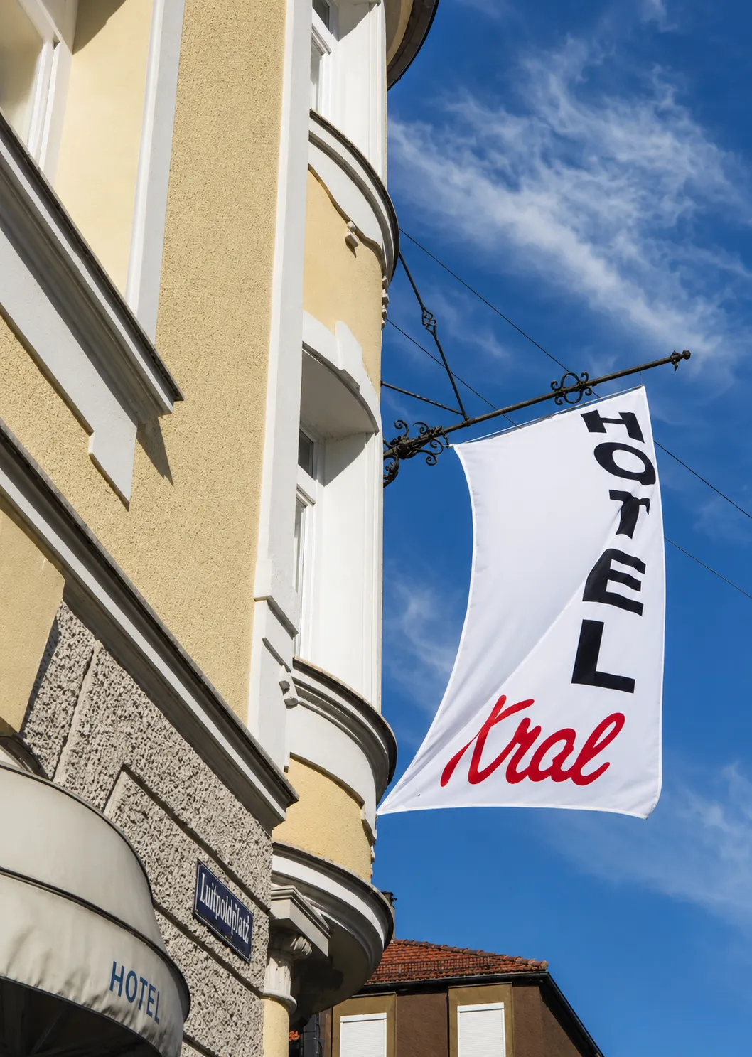 Fahne mit der Aufschrift ‚Hotel Kral‘ an der Ecke eines gelben Gebäudes unter einem blauen Himmel, Straßenschild ‚Luitpoldplatz‘ sichtbar.