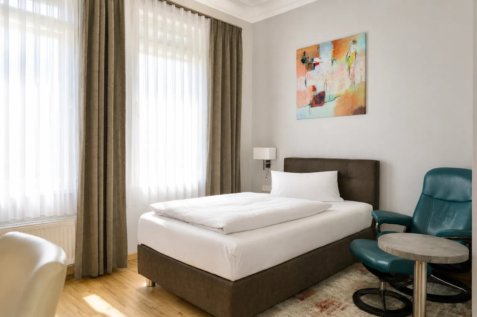Helles Einzelzimmer mit Fenster, braunem Bett, weißer Bettwäsche, blauem Ledersessel, kleinem runden Tisch und abstraktem Gemälde an der Wand.