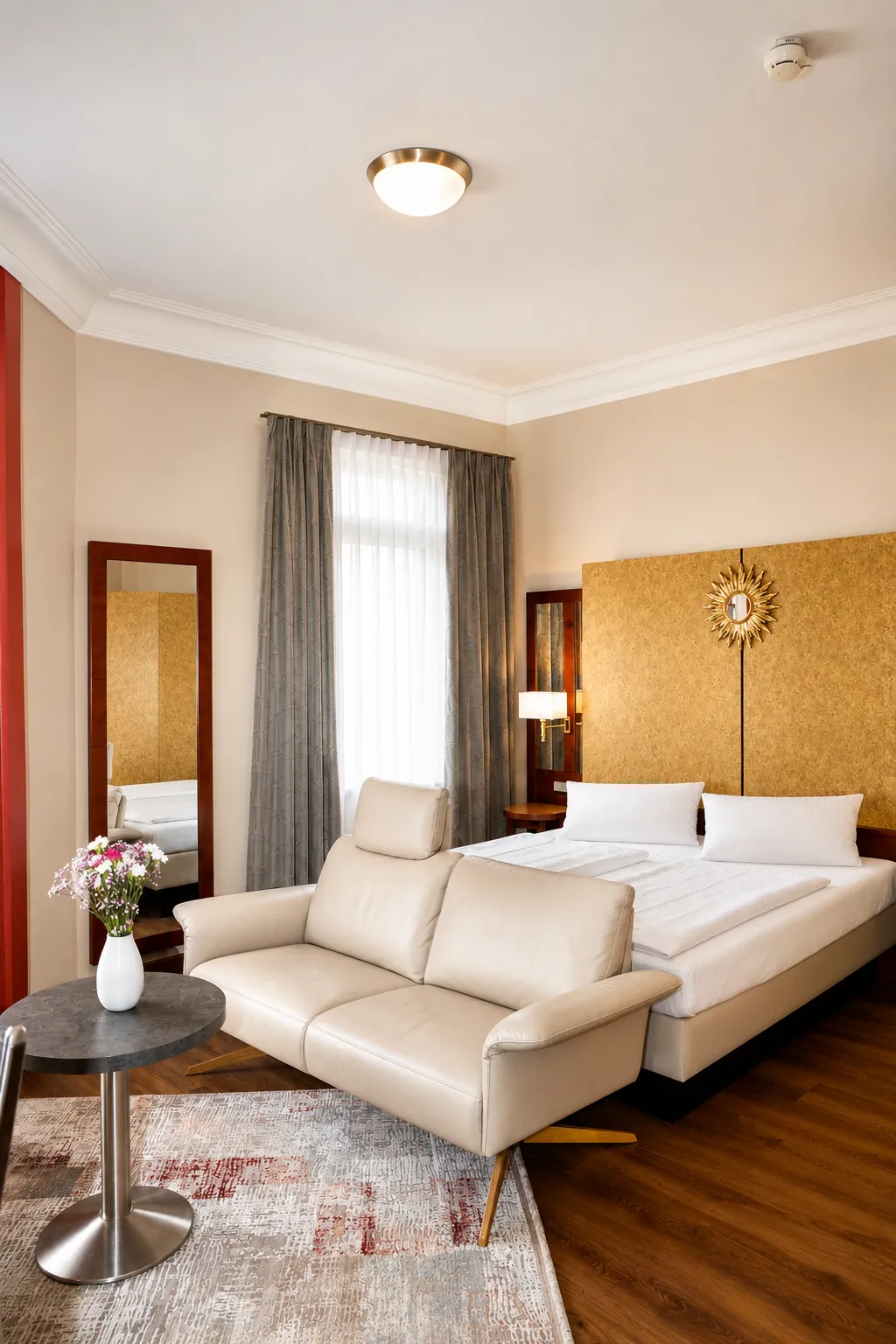 Helles Hotelzimmer mit einem Doppelbett mit weißen Kissen, einem beigen Sofa, einem runden Tisch mit Blumen und einem großen Spiegel neben einem Fenster mit grauen Vorhängen.