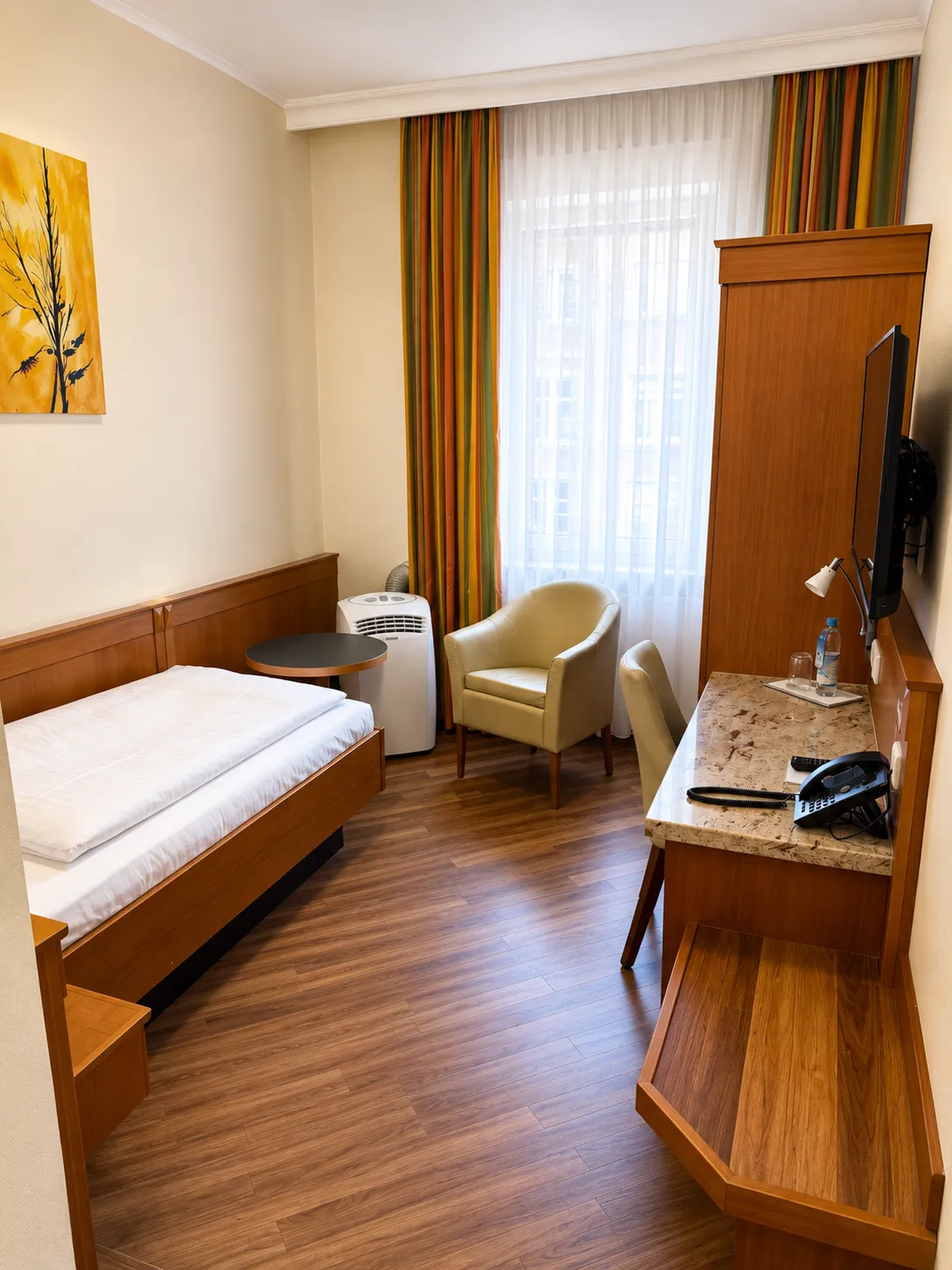 Kleines Gästezimmer mit Einzelbett, Holzfußboden, Schreibtisch mit Telefon, Sessel, rundem Beistelltisch, Gardinen und einer Wanddekoration mit gelbem Blumenmotiv.