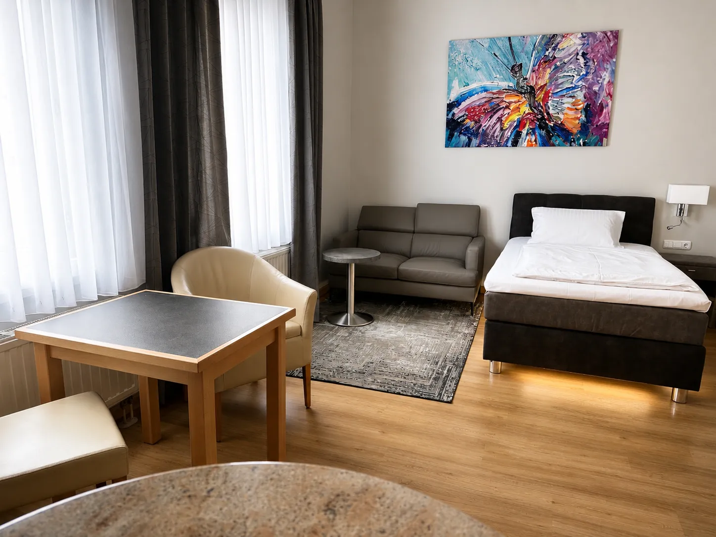 Helles Zimmer mit Holzfußboden, Einzelbett mit weißer Bettwäsche, grauem Sofa, kleinem runden Tisch, einfarbigen Vorhängen und buntem Schmetterlingsgemälde an der Wand.