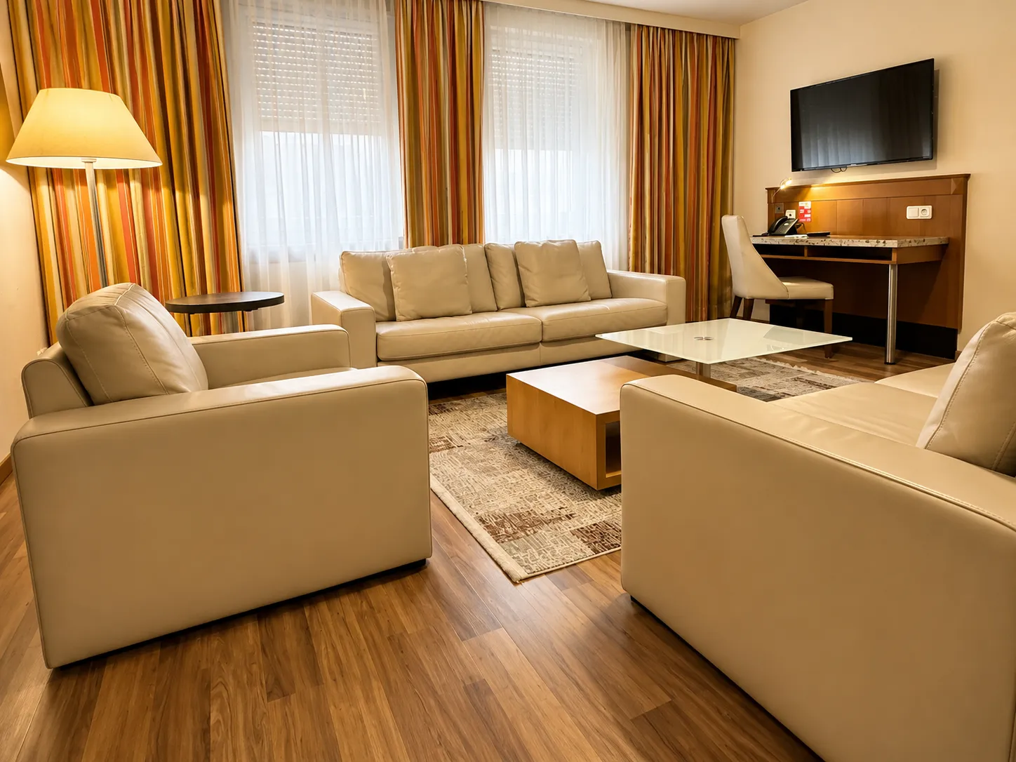 Modern Wohnzimmer mit hellen Ledersofas, einem Glas-Couchtisch, Holzfußboden, Teppich, Stehlampe und einem Wandarbeitsplatz mit Fernseher.