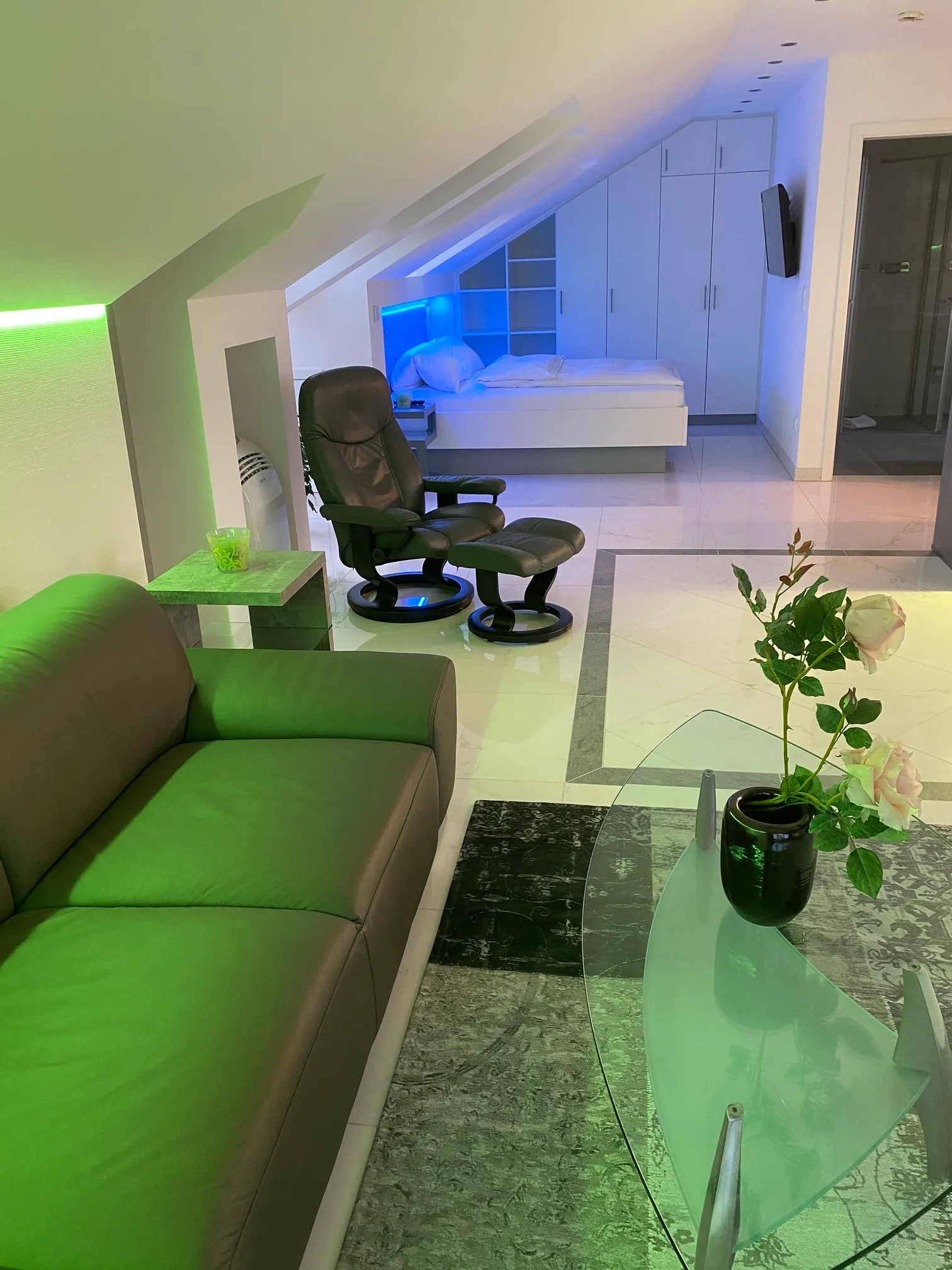Modern attic room with slanted ceiling featuring a green-lit sofa, black leather armchair with matching footrest, glass coffee table with a black vase and flowers, white bed with blue backlight, white closet, and tiled floor.