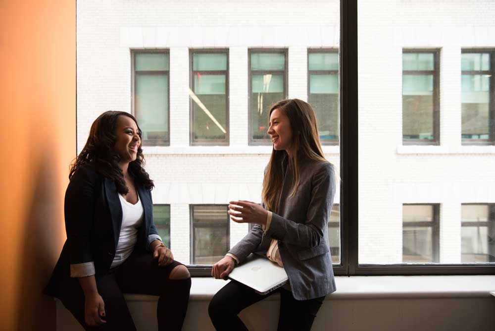 Two women in business attire sitting by a large window, smiling and talking, one holding a laptop.