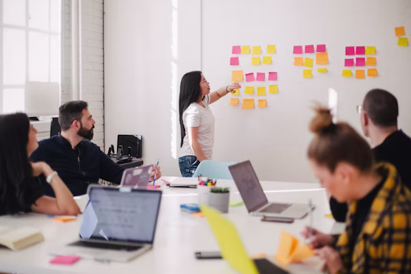 Woman pointing at sticky notes on a wall while colleagues watch and work on laptops around a table.