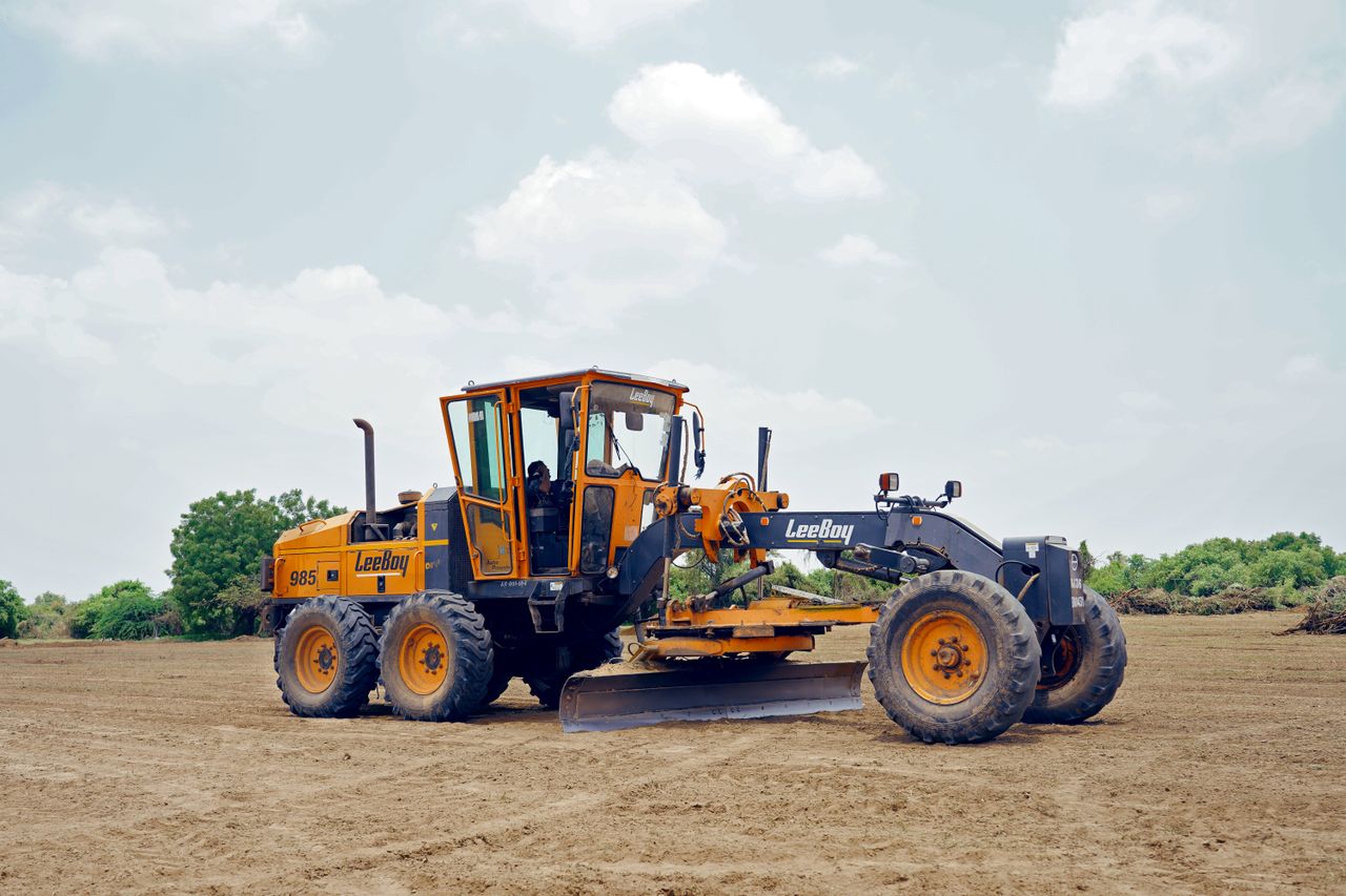 Yellow and black LeeBoy 985 motor grader on a dirt field with scattered green bushes.