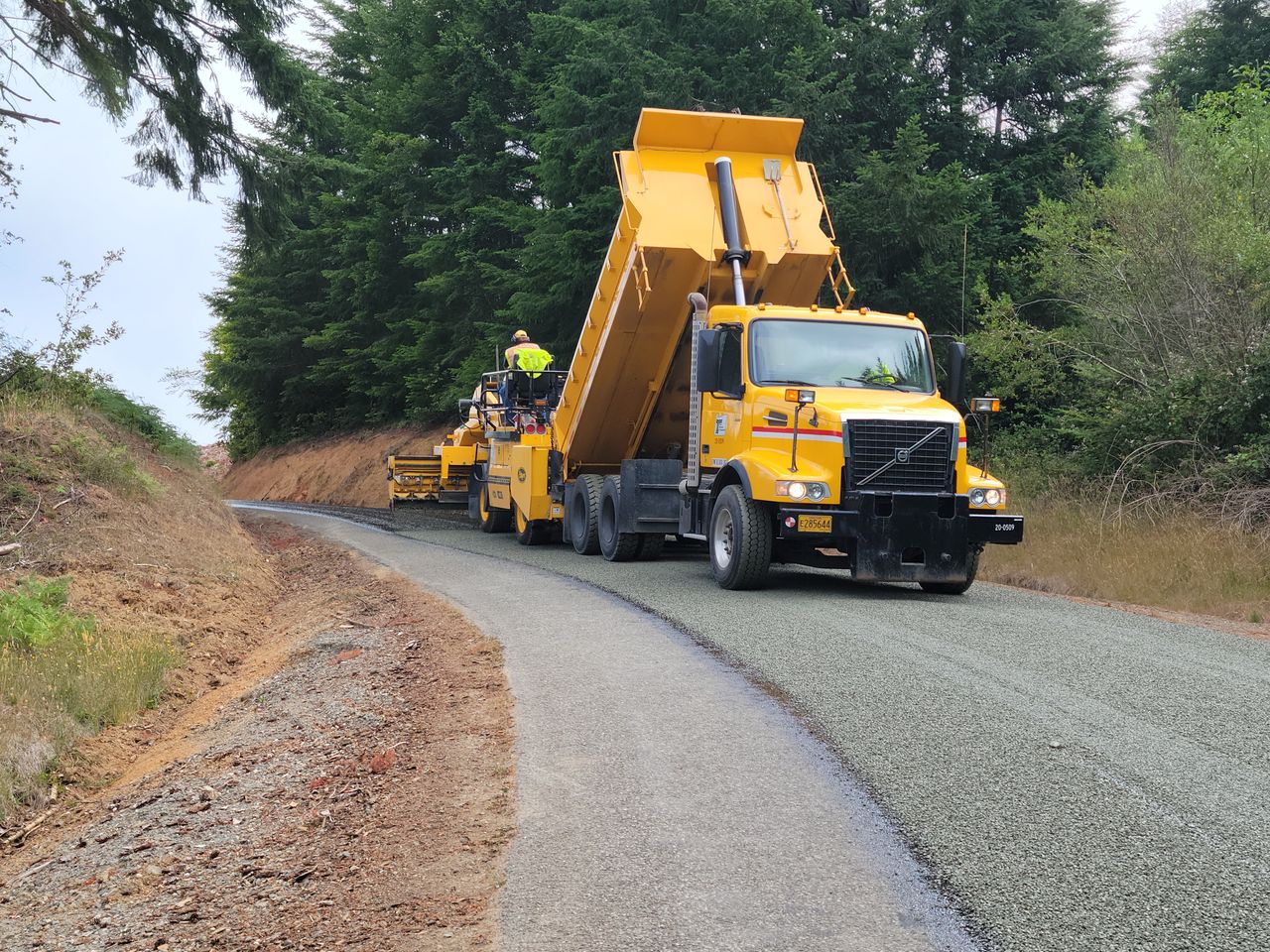 Yellow dump truck unloading gravel for road paving in a forested area with workers operating machinery.