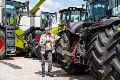 Man in a cap inspecting large tractors parked on a gravel lot under a blue sky.