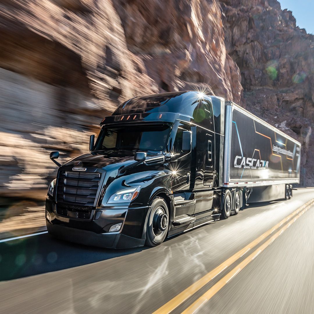 Sleek black Cascadia semi-truck driving fast on a road through rocky terrain under clear sky.