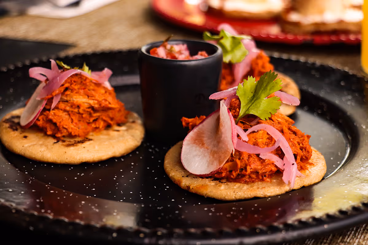 Three panuchos topped with pulled meat, pickled onions, radish slices, and cilantro on a black plate.