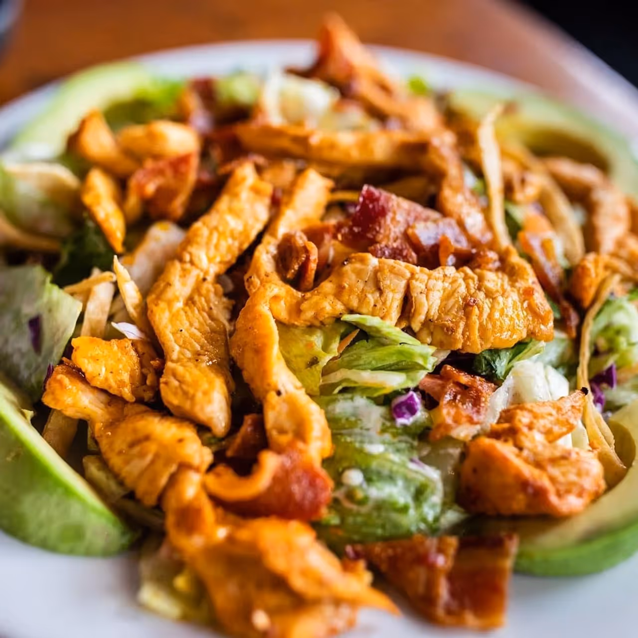 Close-up of grilled chicken strips on a fresh salad with lettuce, avocado slices, and bacon bits.