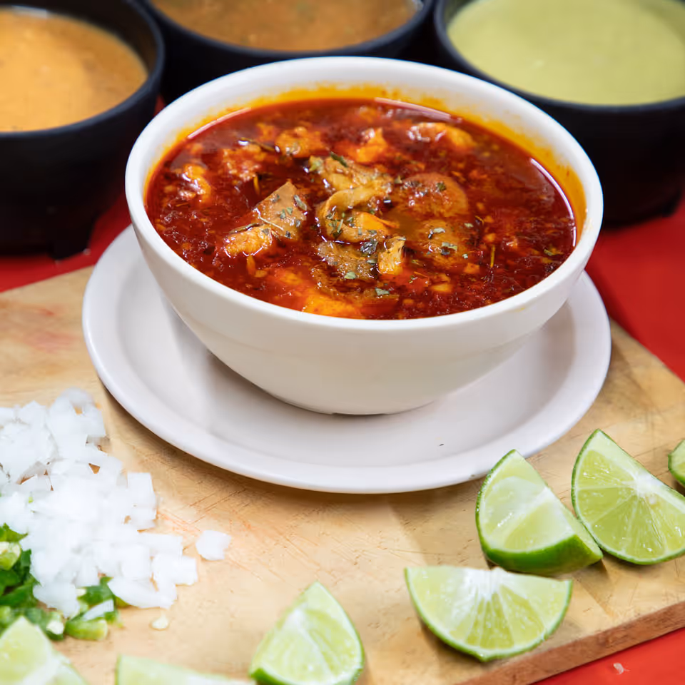 Bowl of spicy red menudo soup with chopped onions, lime wedges, and sauces on a wooden board.