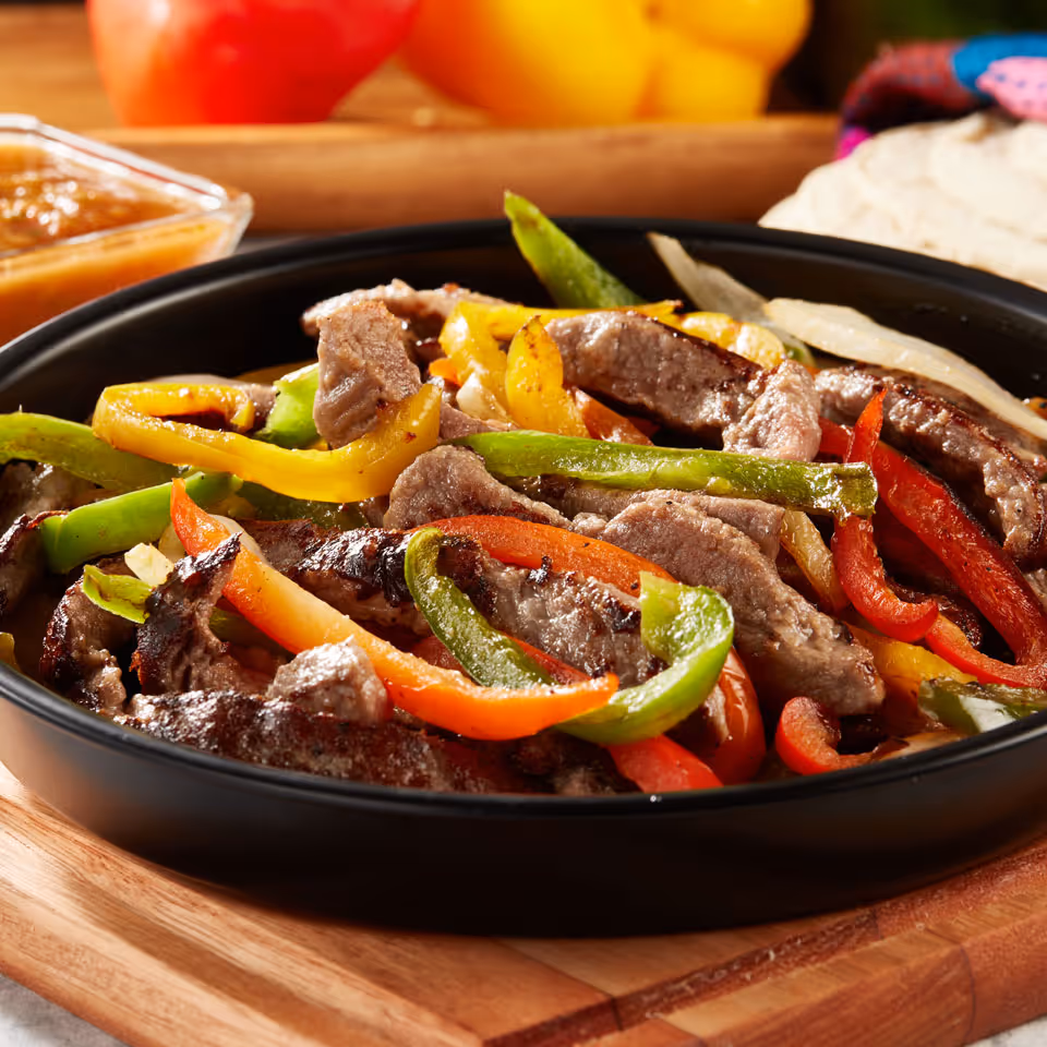 Skillet of cooked beef strips with red, yellow, and green bell peppers and onions on a wooden board.