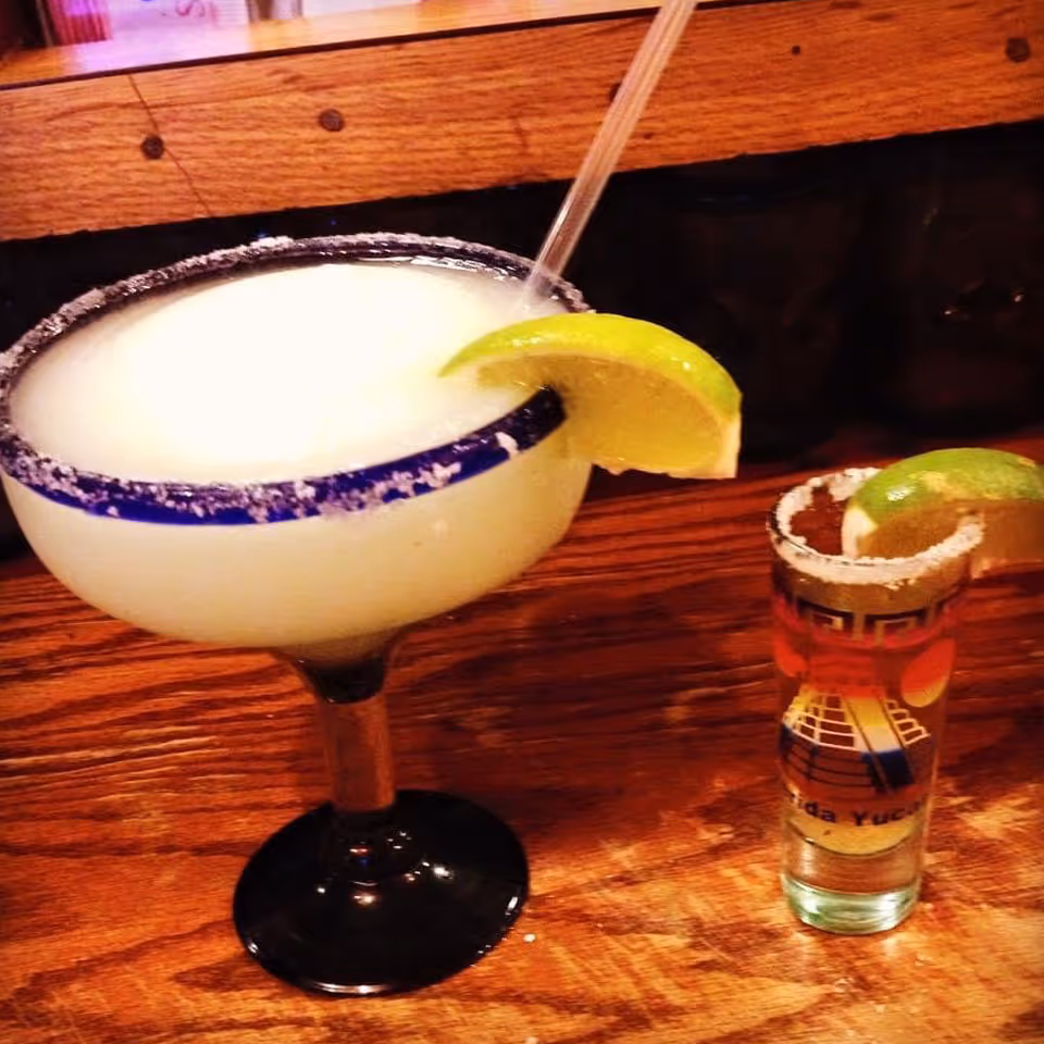 Frozen margarita with lime wedge and a shot glass with lime on a wooden bar surface.