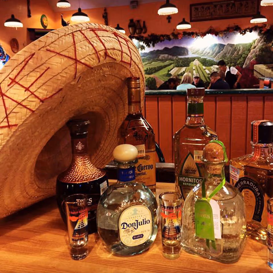 Table with bottles of tequila, shot glasses, and a large woven sombrero hat in a Mexican restaurant.