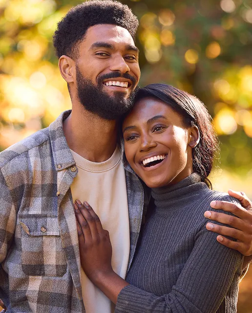 Smiling couple embracing outdoors with blurred autumn foliage in the background.