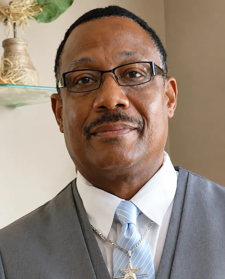Man wearing glasses, grey suit, white shirt, and a light blue striped tie with a star pendant necklace.