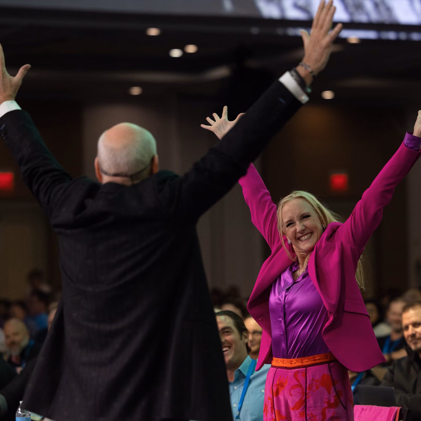 Smiling woman in pink raises her arms in celebration facing a man in a dark jacket with arms raised.