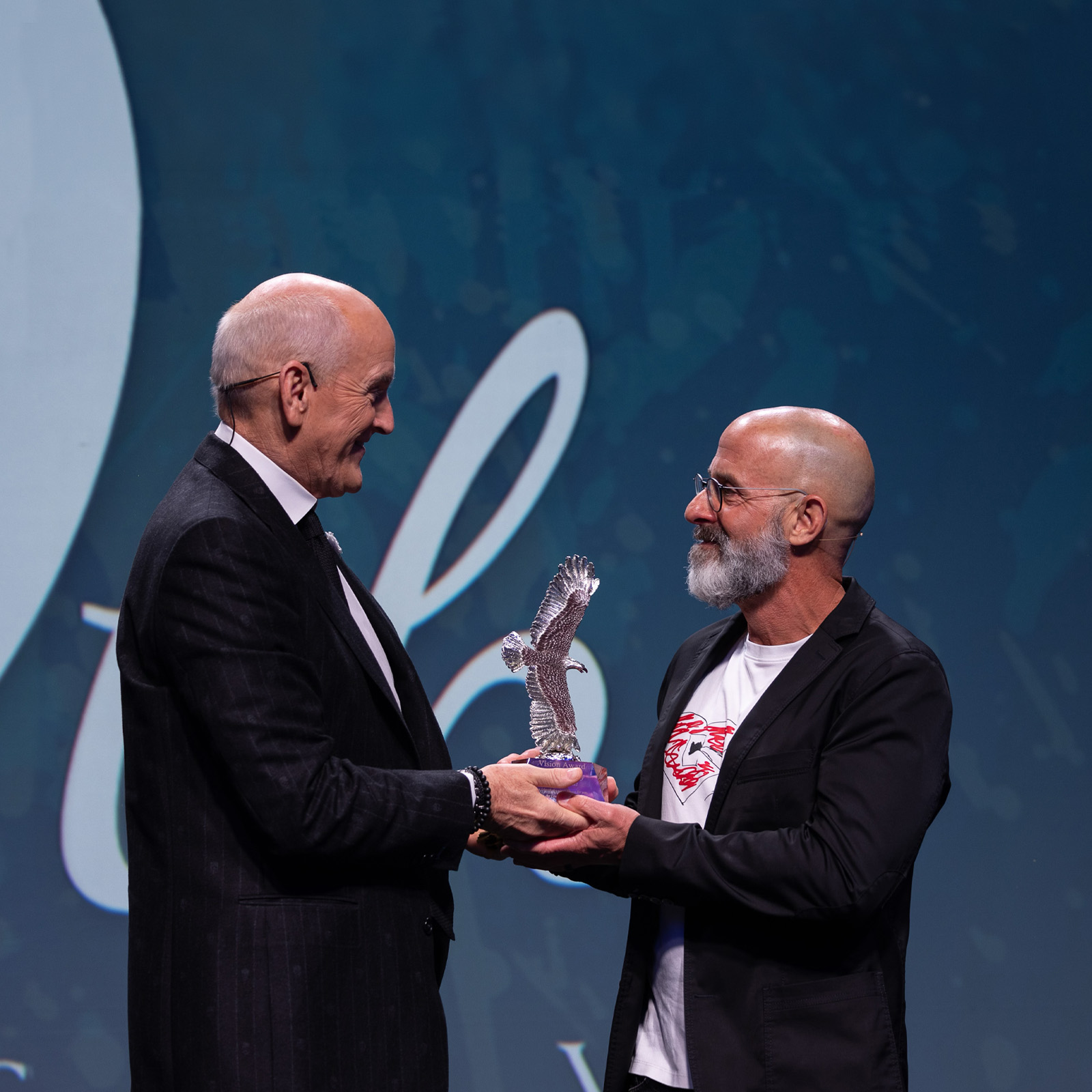 Two men smiling as one presents an eagle-shaped glass trophy to the other on stage.
