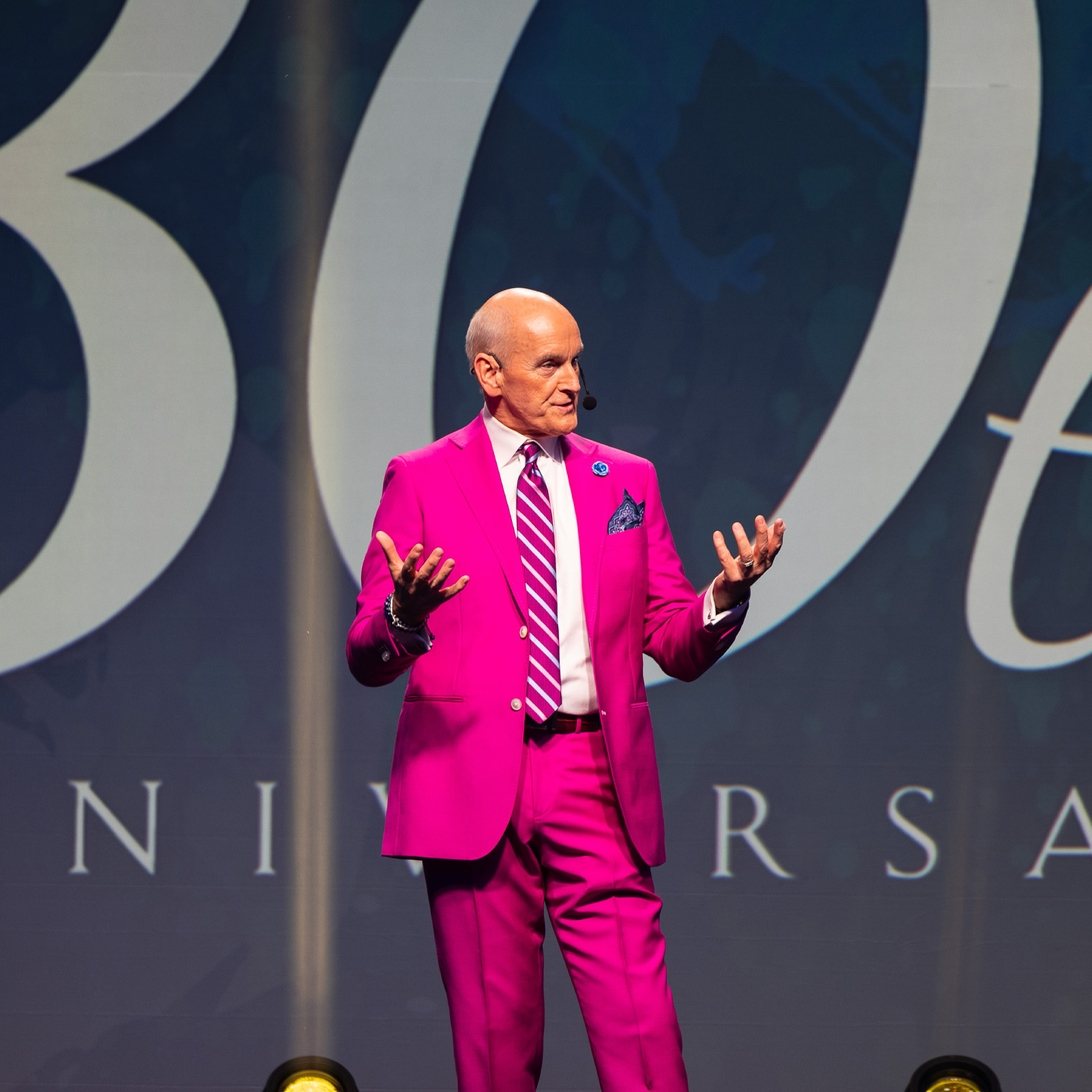 Man in bright pink suit speaking on stage in front of large backdrop with text.