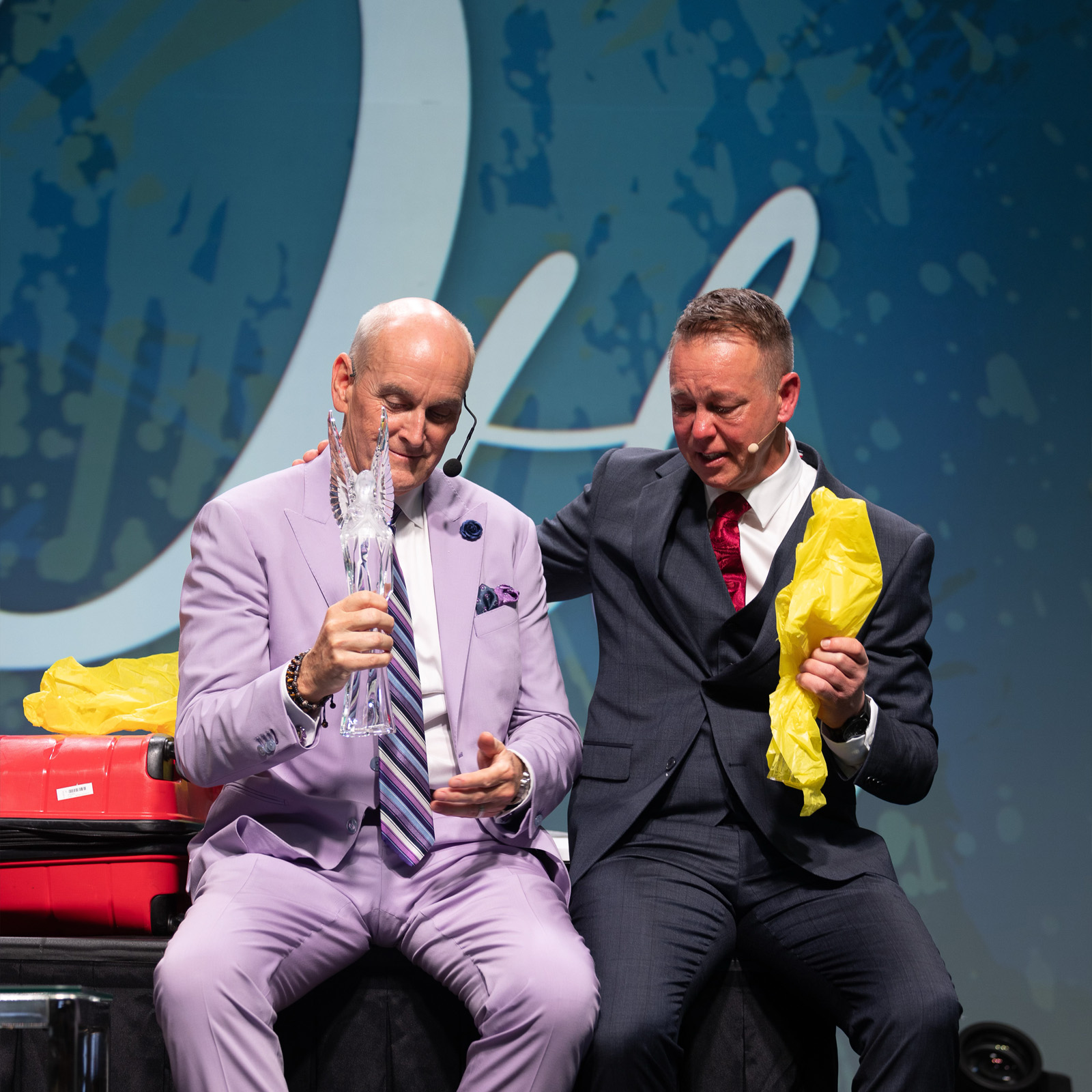 Two men in suits on stage, one holding a clear winged trophy, the other holding yellow paper.