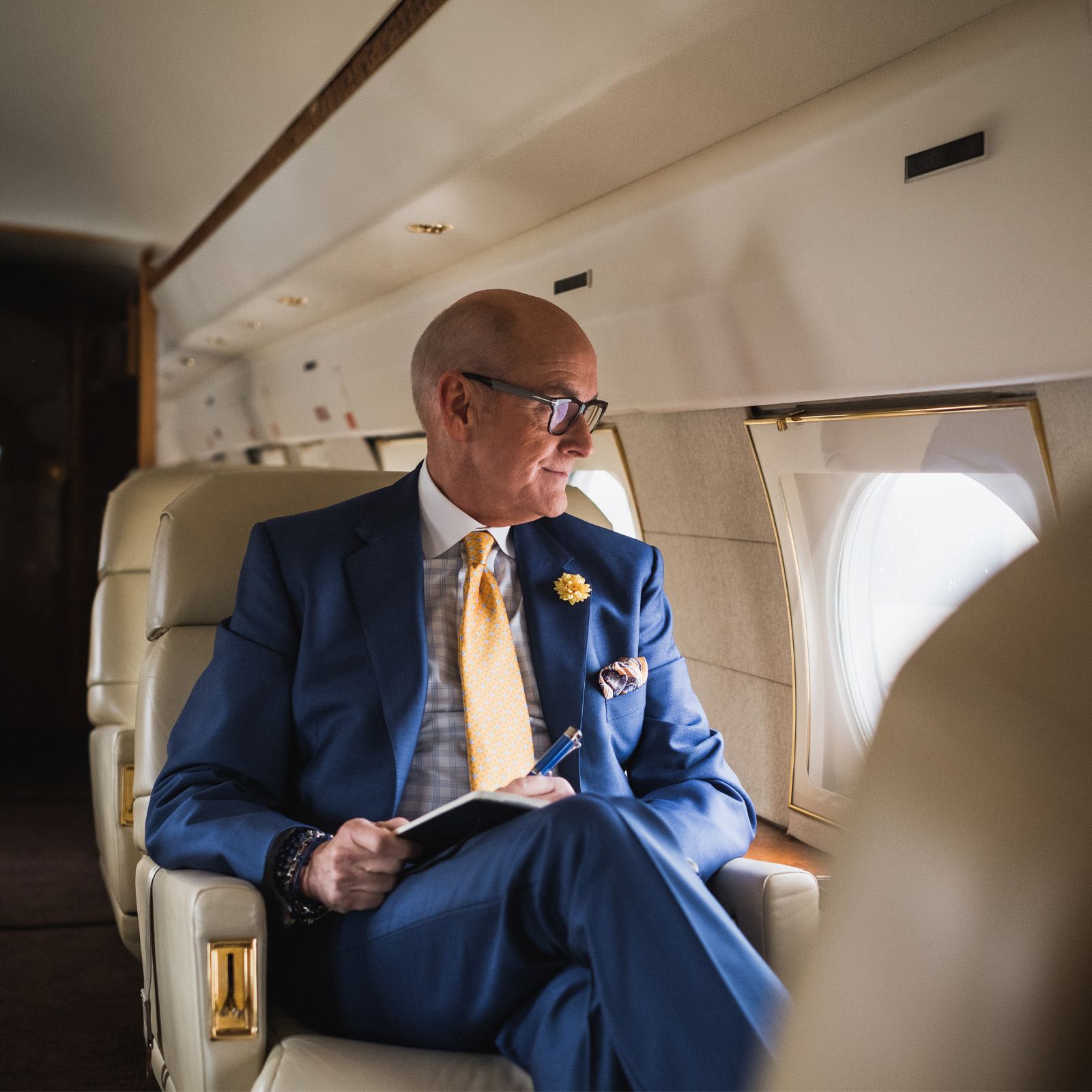 Man in blue suit and yellow tie sitting in private jet seat, looking out the window while holding a notebook.