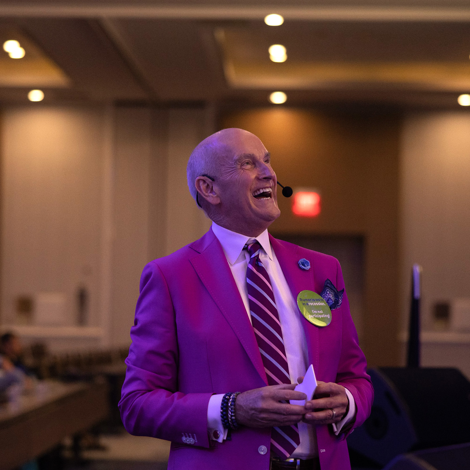 Smiling bald man in purple suit and striped tie speaking on stage with a microphone headset.