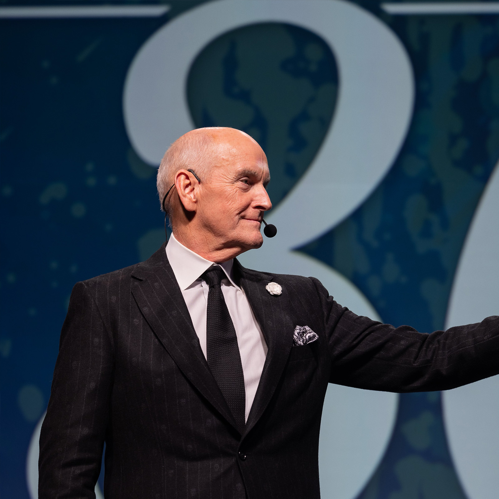 Bald man in a suit speaking on stage with a headset microphone and large number 30 behind him.