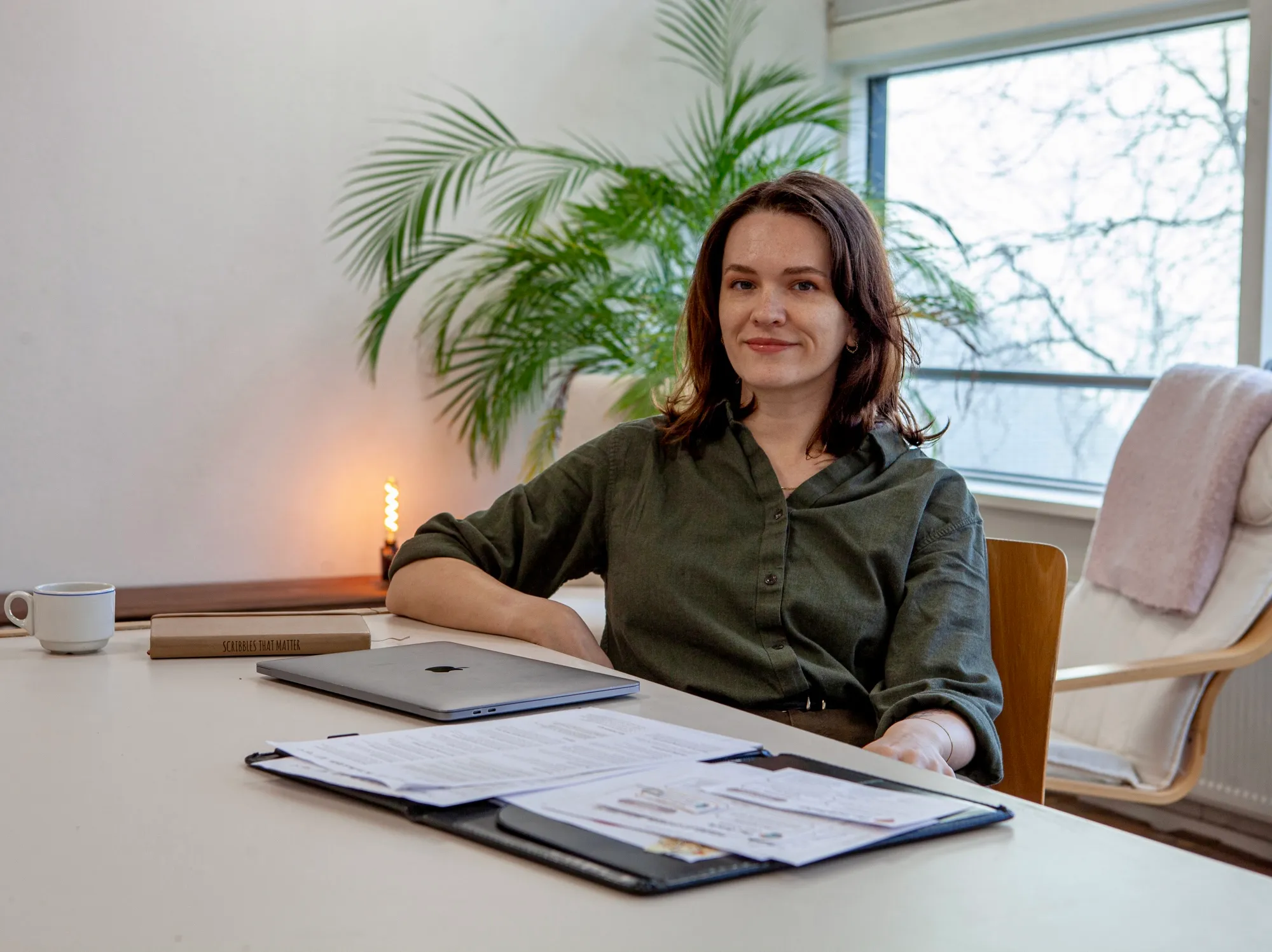 Woman sitting at a desk with a laptop, documents, and a cup, with a plant and window in the background.