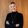 Smiling woman with arms crossed wearing a black turtleneck standing in front of a wooden panel wall.