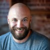 Smiling bald man with a beard wearing a blue shirt against a blurred brown background.