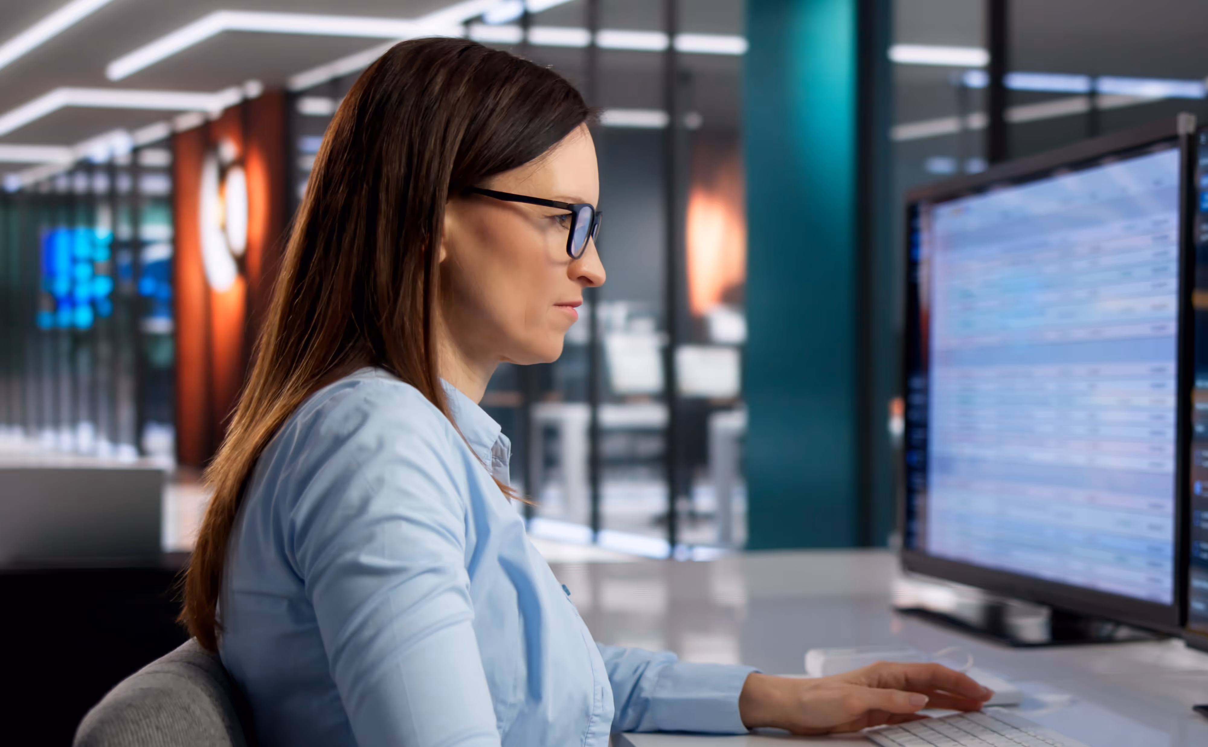 Woman with glasses in blue shirt focused on computer screen displaying data in modern office.