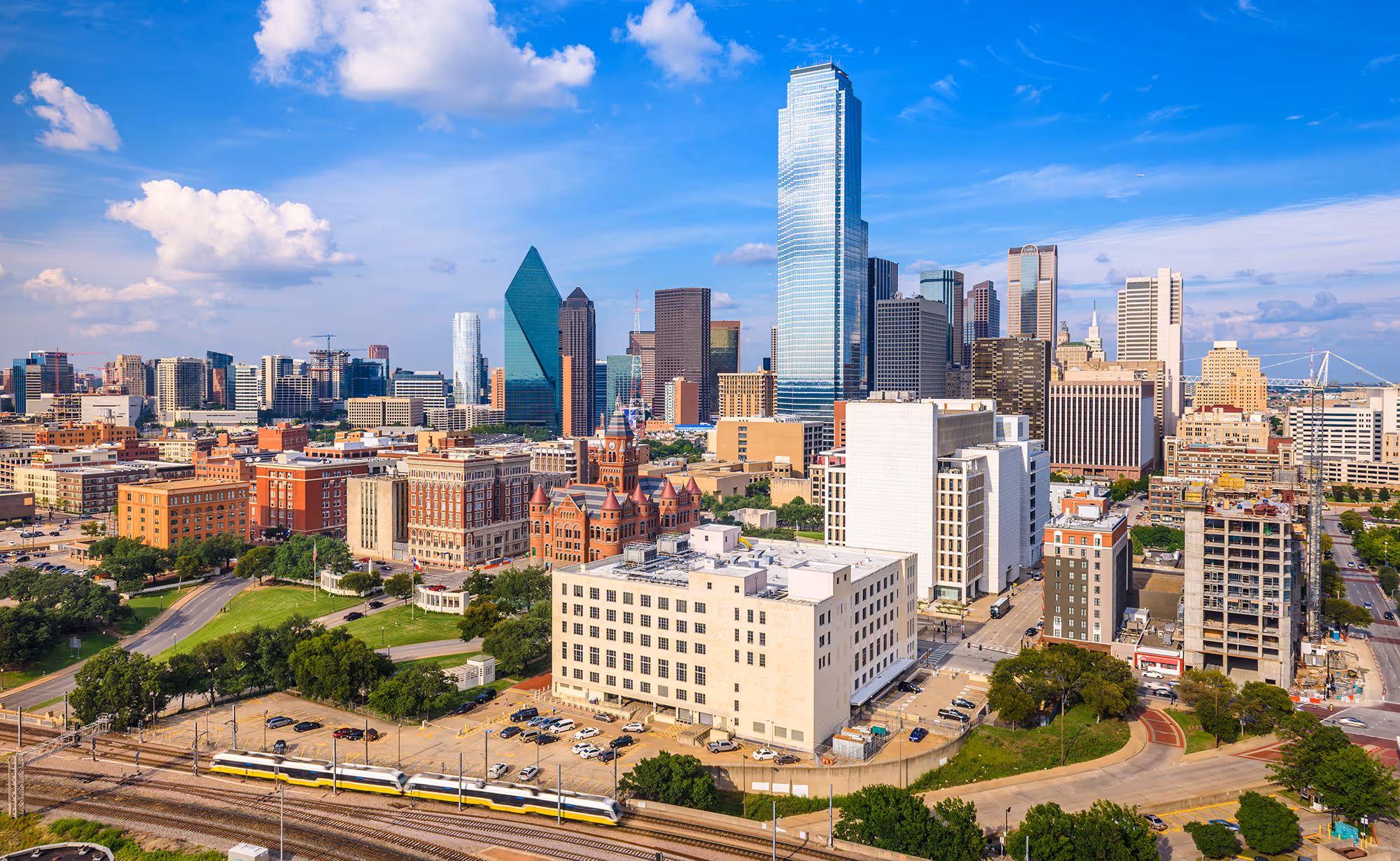 Downtown Dallas skyline with tall buildings, a train passing in the foreground, and a blue sky with clouds.
