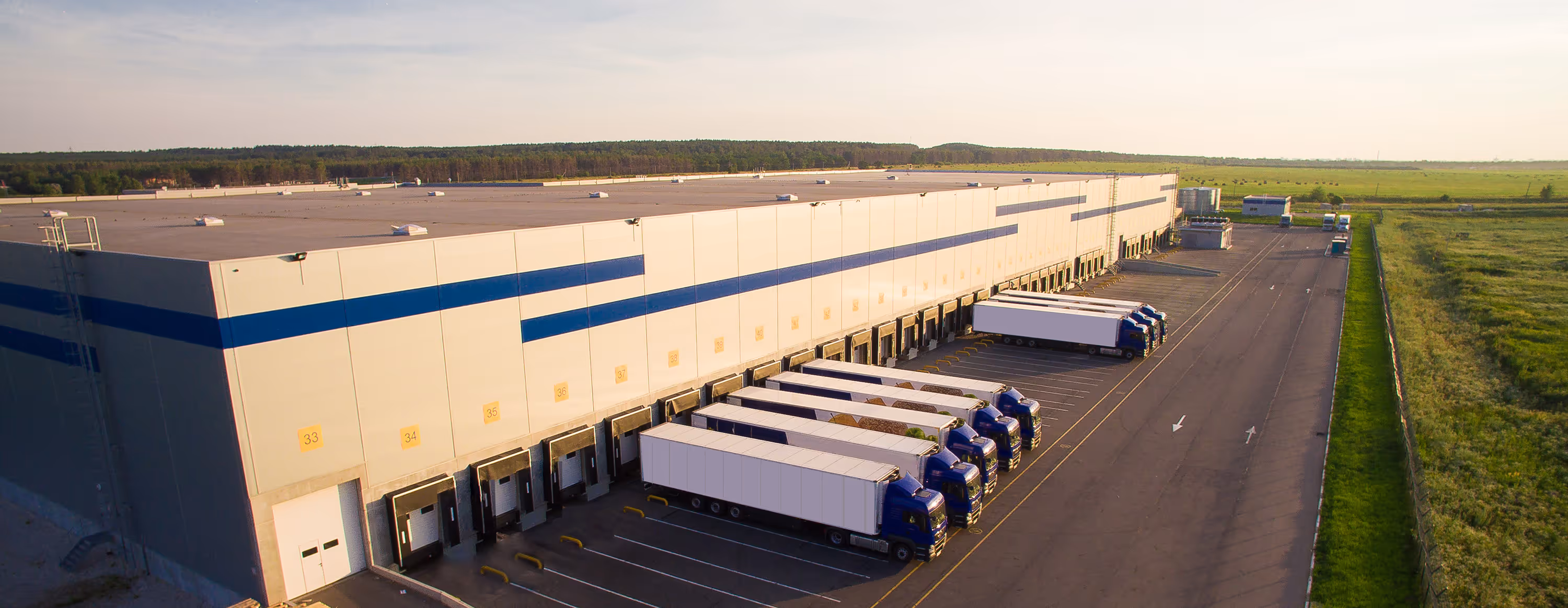 Several semi trucks backed into loading docks of a large warehouse with green fields nearby.