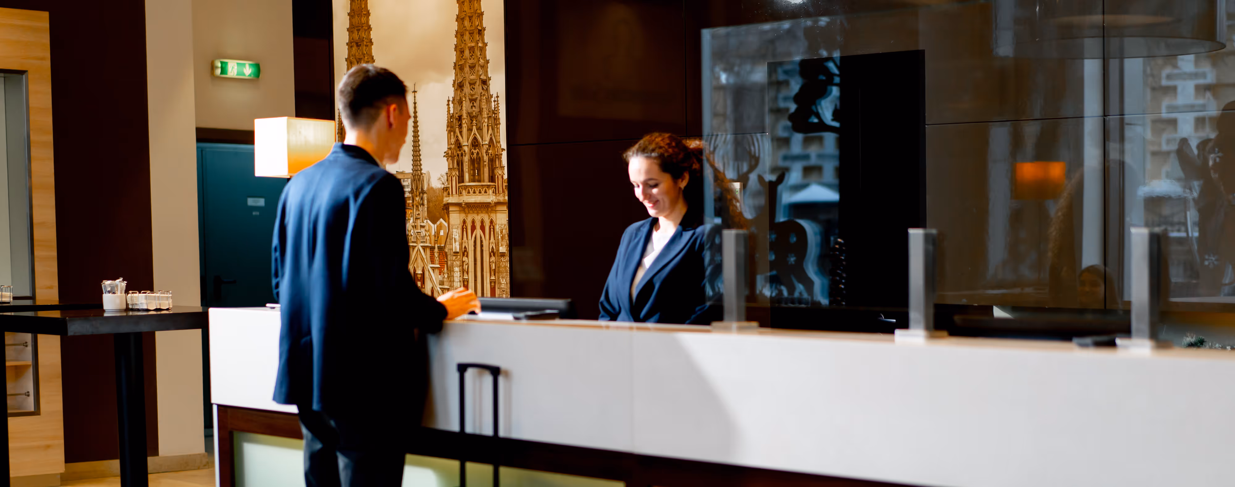 Man with suitcase at hotel reception desk speaking to smiling female receptionist behind a protective screen.
