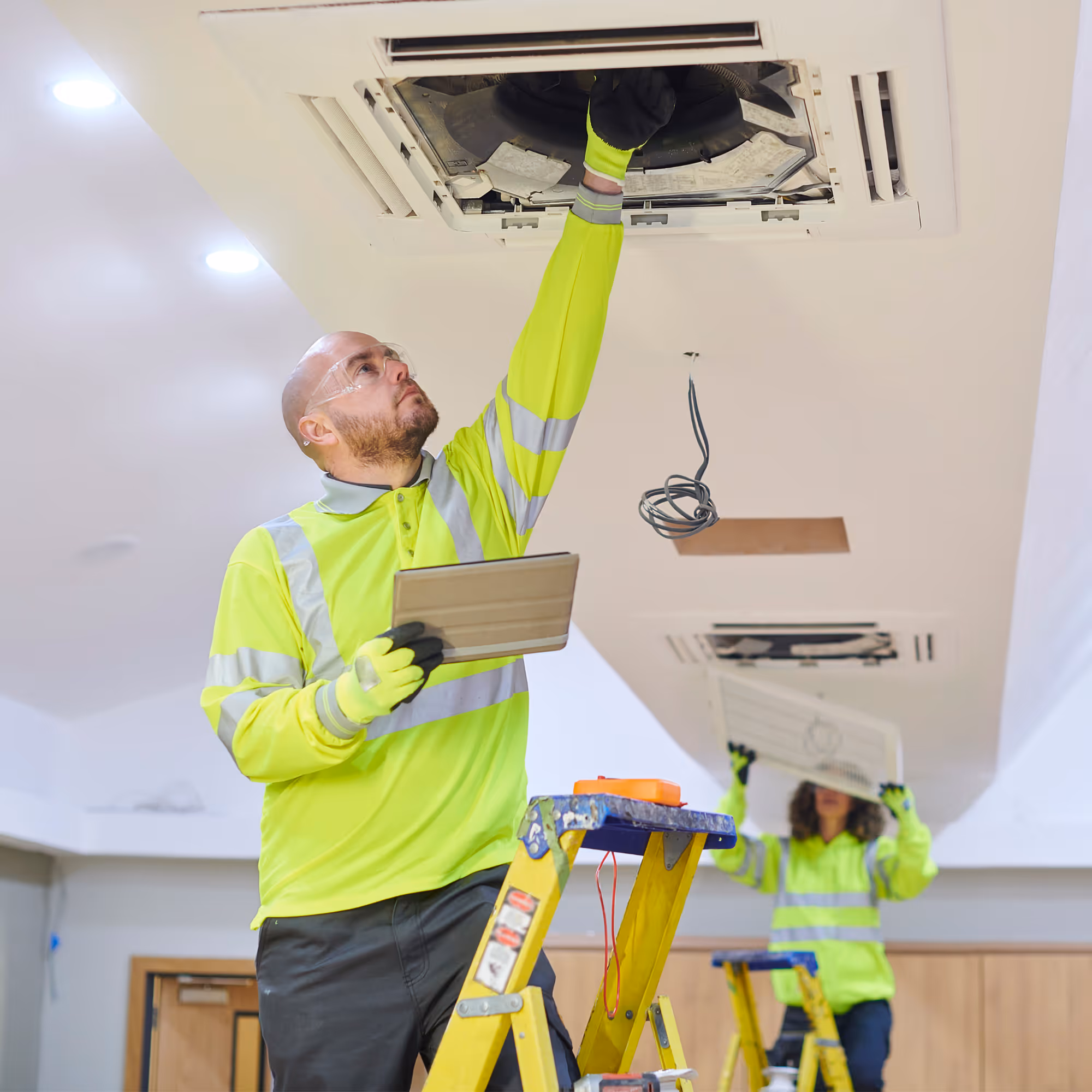 Two technicians in neon yellow safety gear repairing ceiling air conditioning units, one standing on a ladder inspecting the unit while holding a tablet.