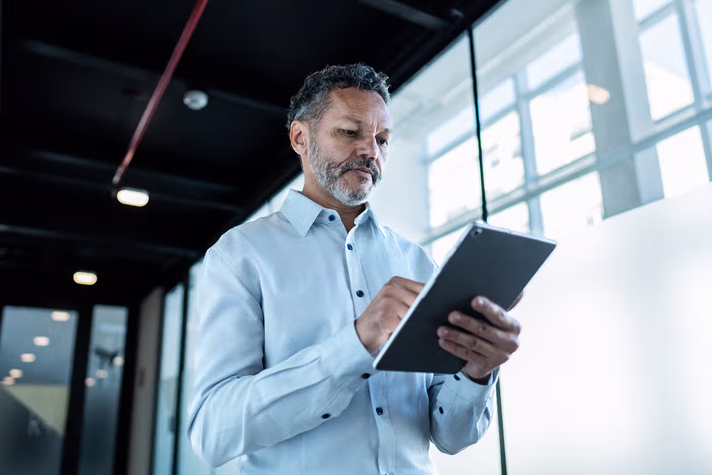 Man with gray beard and light blue shirt using a tablet in a modern office with large windows.