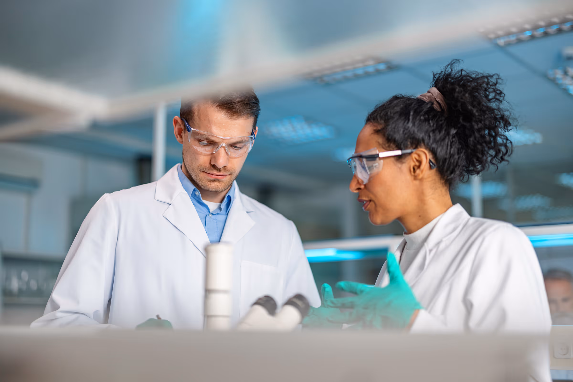 Two scientists wearing lab coats and safety goggles discussing in a laboratory.