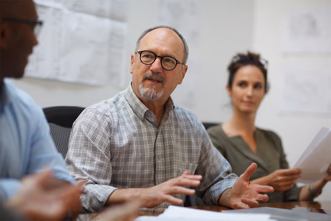 Middle-aged man with glasses speaking during a meeting with two colleagues in an office.