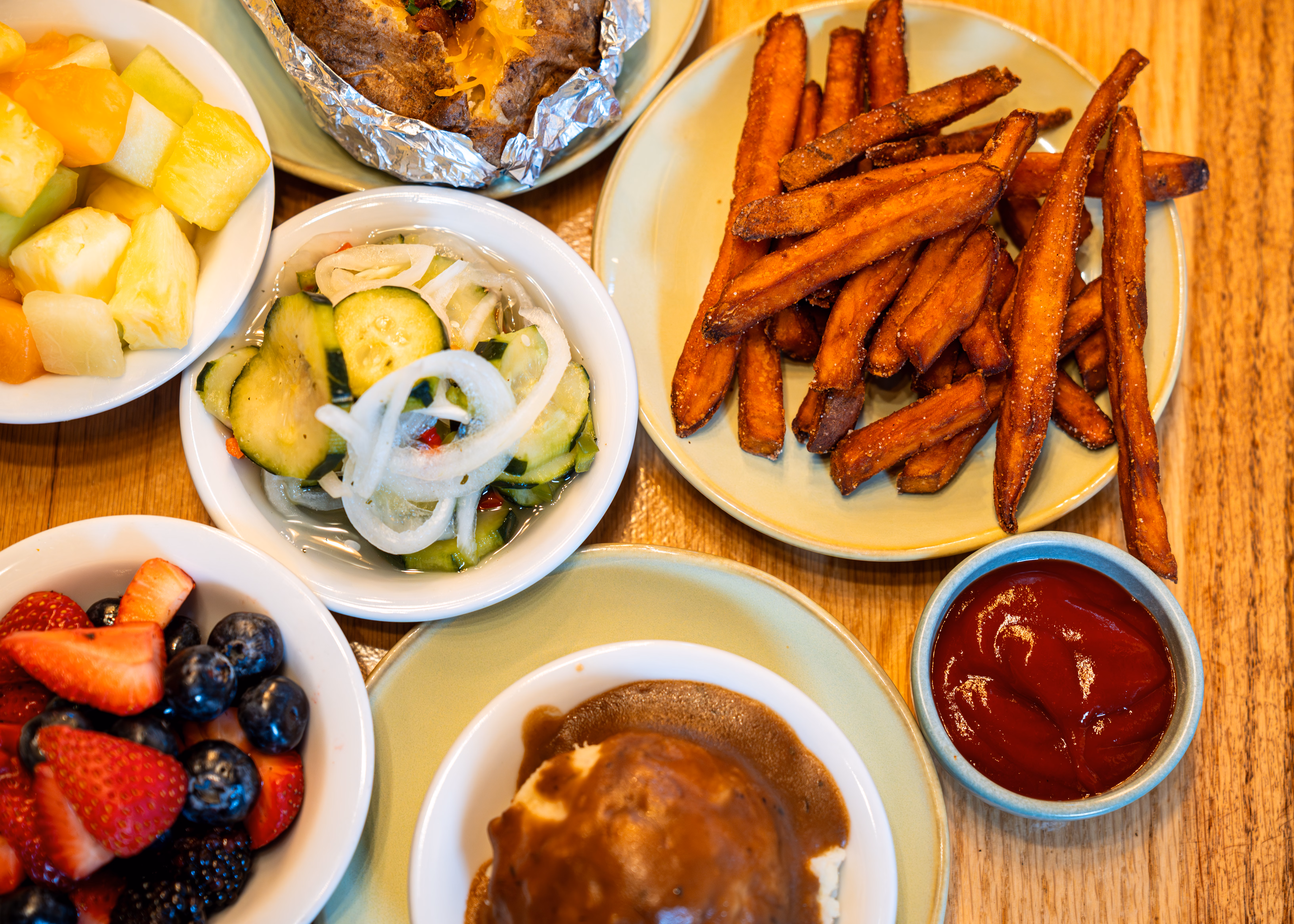 A colorful assortment of dishes on a wooden table: sweet potato fries with ketchup, fresh fruit, cucumber salad, and mashed potatoes with gravy.