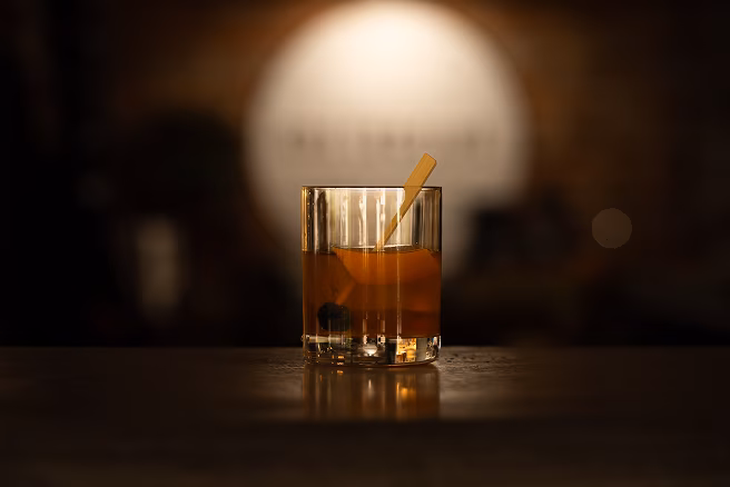 Cut-crystal whiskey tumbler with amber bourbon and ice on a wooden bar, beside a metal jigger; warm, dim lighting creates a cozy mood.