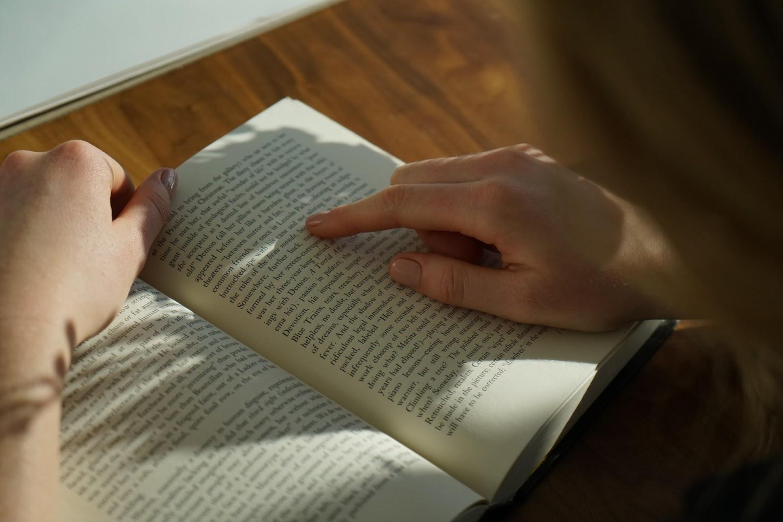 La imagen muestra las manos de una mujer sobre un libro. Con el dedo índice de la mano derecha sigue la lectura del libro. En la esquina superior derecha de la imagen, se puede ver el cabello rubio desenfocado de la mujer.