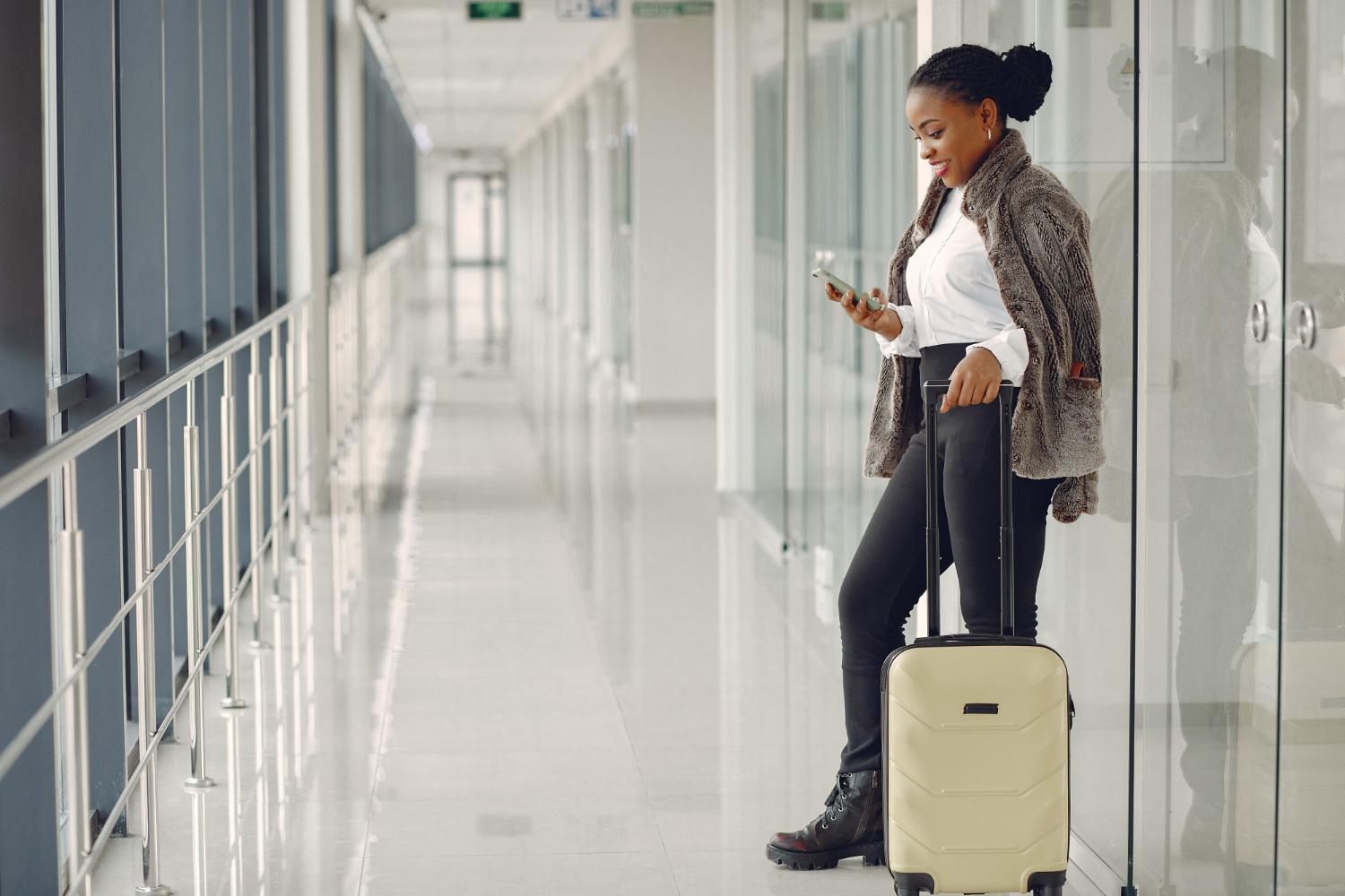 La imagen muestra a una mujer negra que está de lado. Ella está en la esquina derecha de la foto y sostiene una bolsa de viaje con ruedas color crema.