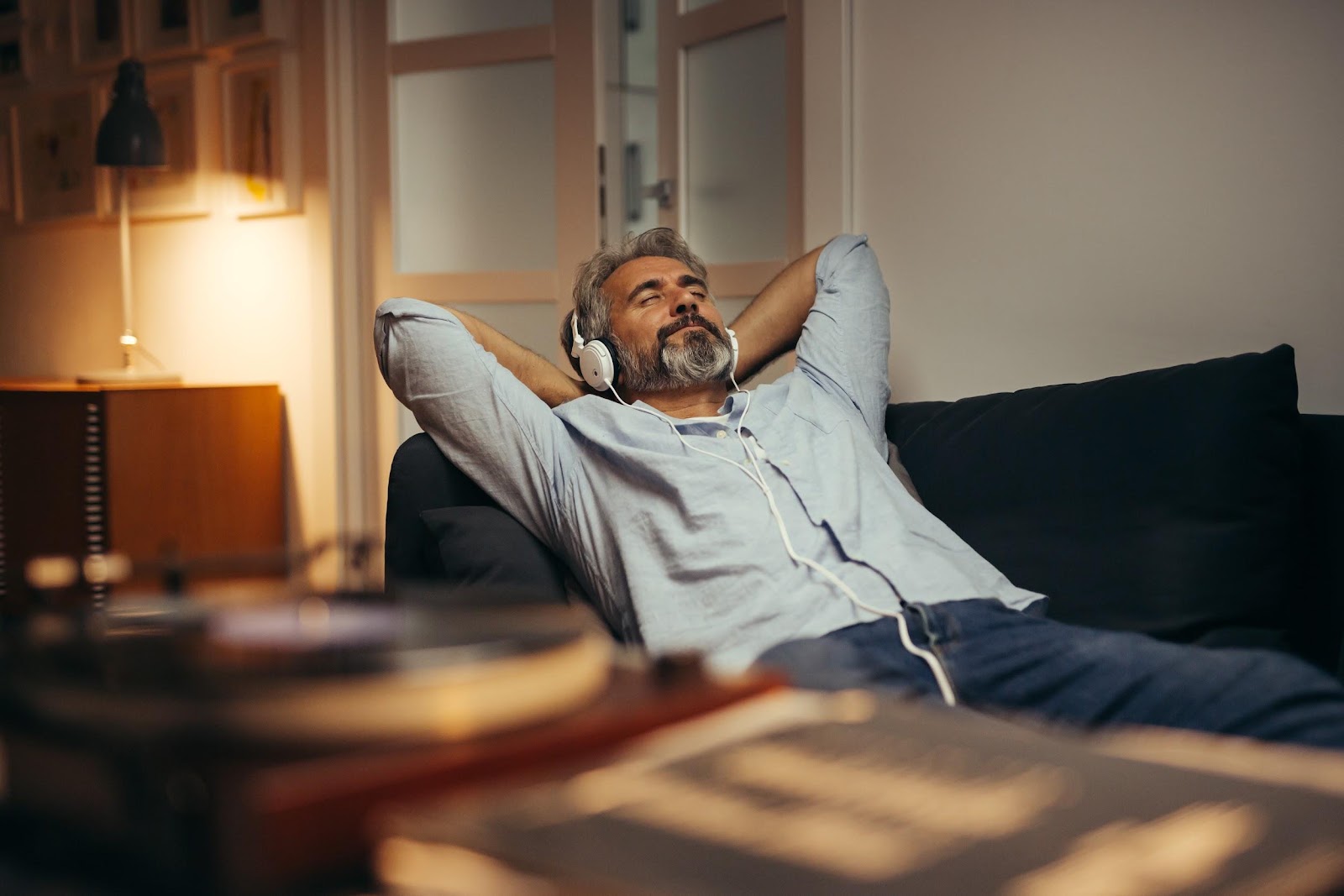 Hombre canoso y barbudo acostado en un sofá negro, con los brazos detrás de la cabeza y escuchando música con audífonos.