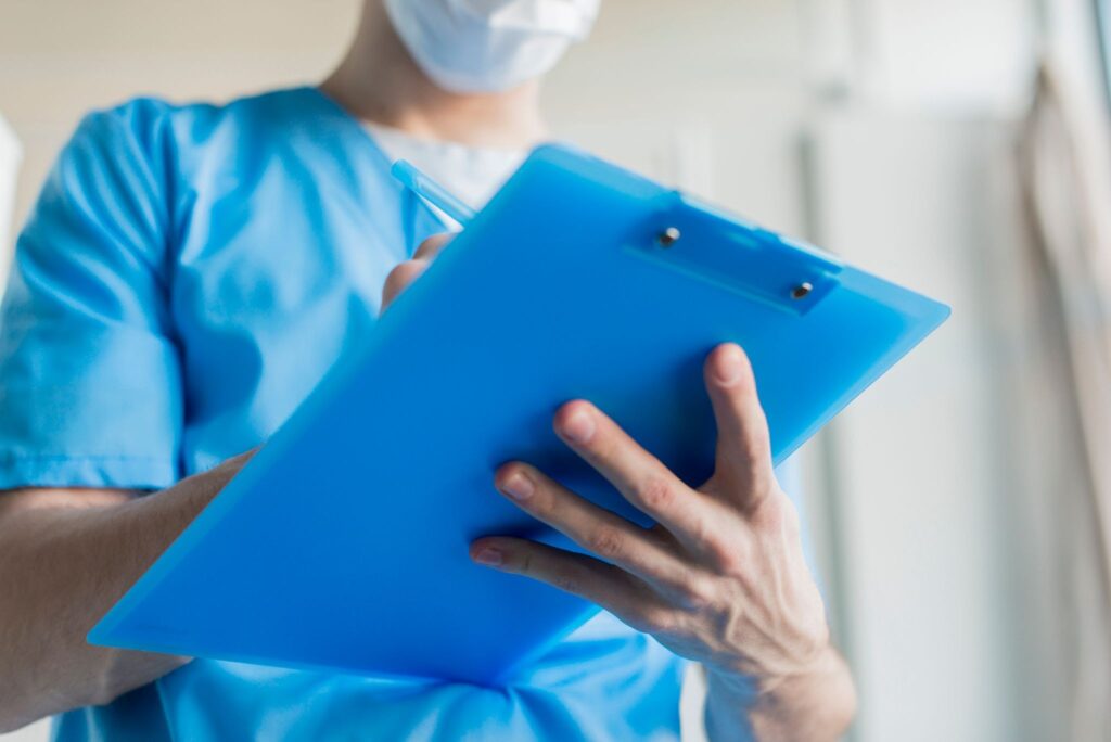 La imagen muestra el torso de un hombre con ropa de color azul, el cual es el uniforme usado por los profesionales de la enfermería. En sus manos hay un portapapeles azul en el que toma notas.
